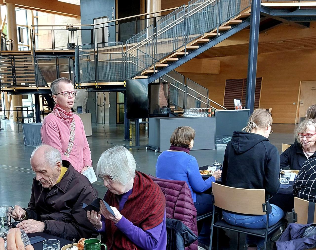 Kuvan etualalla kaksi ihmistä ruokailemassa ja heidän takanaan puheenjohtaja Aila pitämässä puhetta. Taustalla näkyy Metsähallista nousevat portaat ylös kohti konserttisalin parvea.